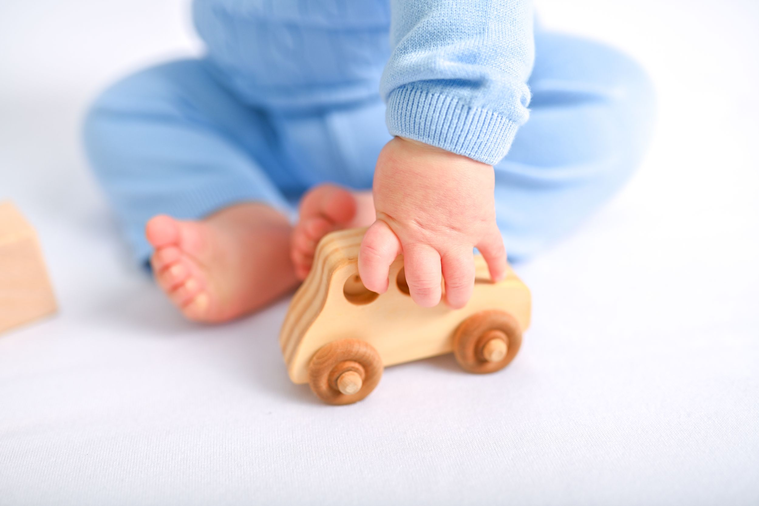 Baby wearing light blue clothing reaching for wooden toy car on white surface.