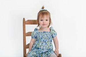 Child in blue floral dress sitting on wooden chair against white backdrop.