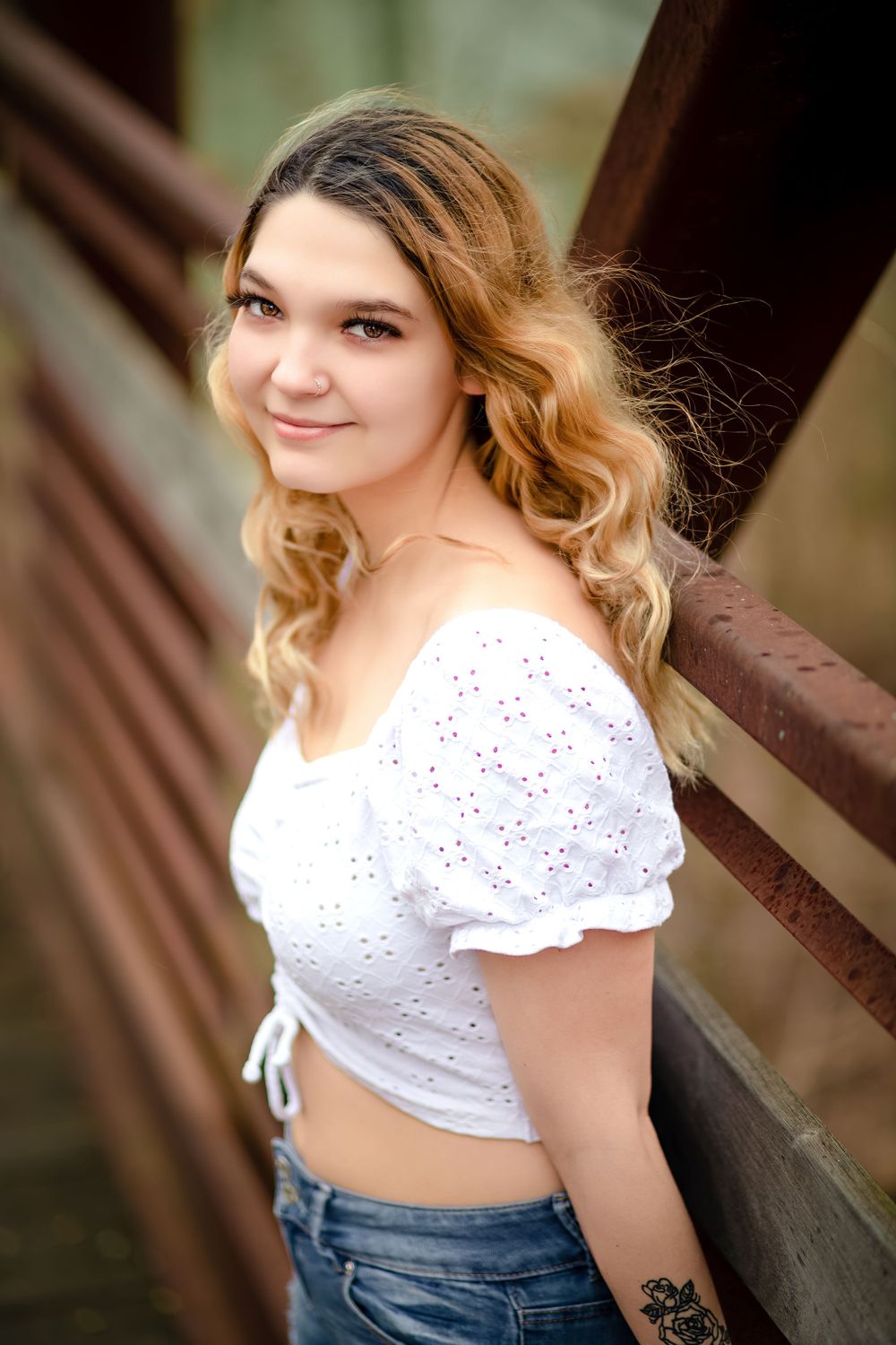 A portrait of someone in a white eyelet crop top and jeans standing against a wooden railing outdoors.