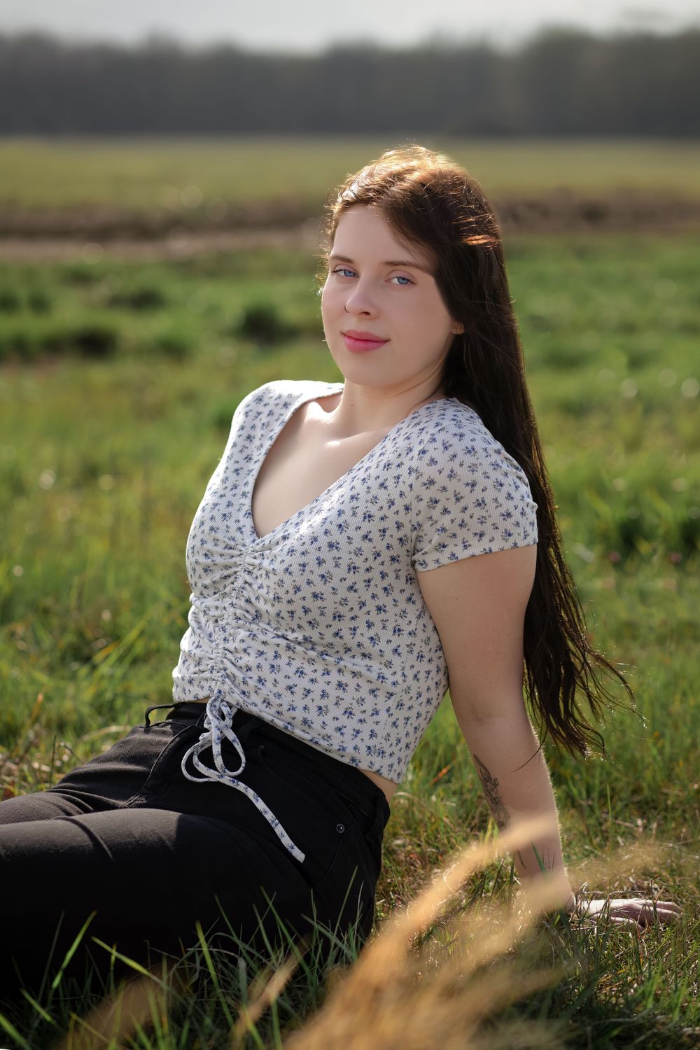 A young person in a floral top and black skirt sits in a sunlit meadow during golden hour.