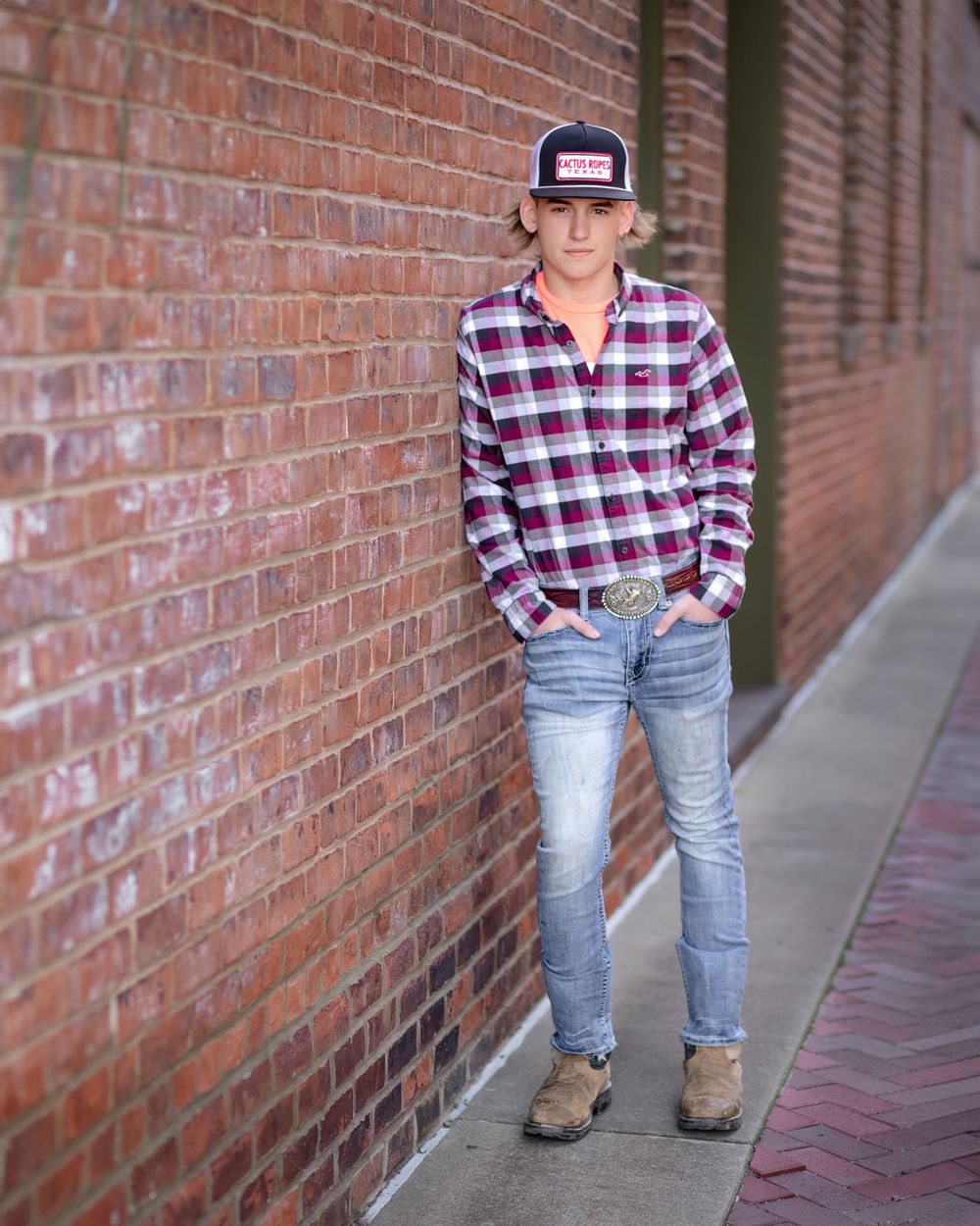 Person in plaid shirt and baseball cap leans against brick wall in casual urban portrait.