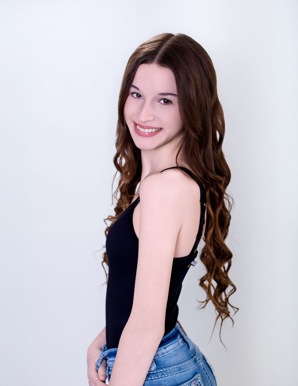 Young woman in a black tank top and jeans poses sideways against a white background with long wavy brown hair.