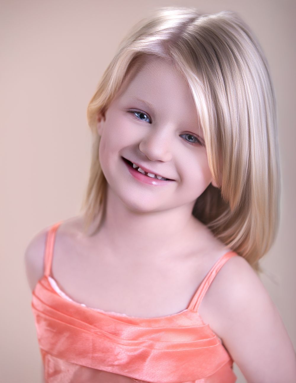 Portrait of a young child with straight blonde hair wearing a peach-colored top smiling warmly at the camera.
