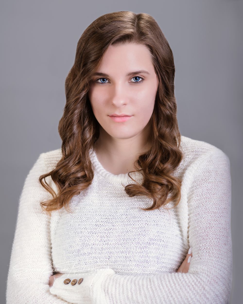 Portrait of a person with long wavy brown hair wearing a white knit sweater against a gray background.