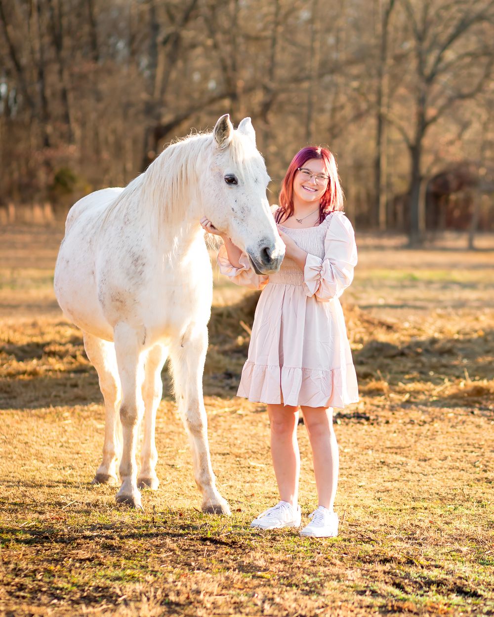 Beautiful white horse and young woman in a pink dress stand together in a sunlit autumn field.