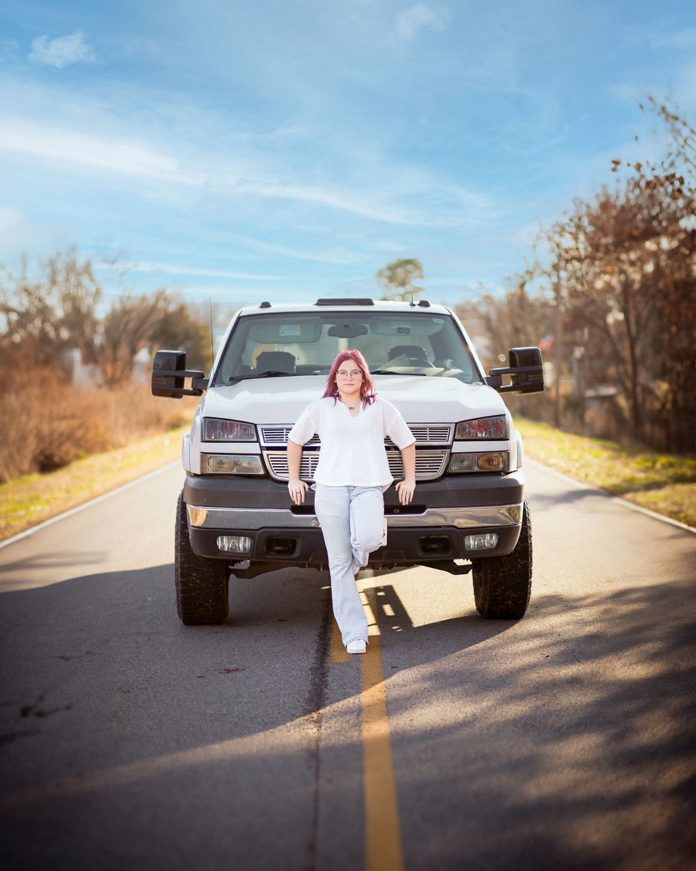 Person in white outfit poses confidently in front of lifted pickup truck on rural road.