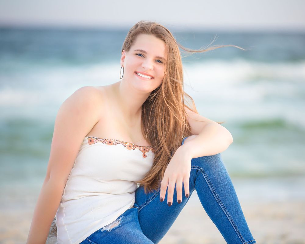 Portrait of someone in white top and jeans sitting on beach with ocean waves in background.