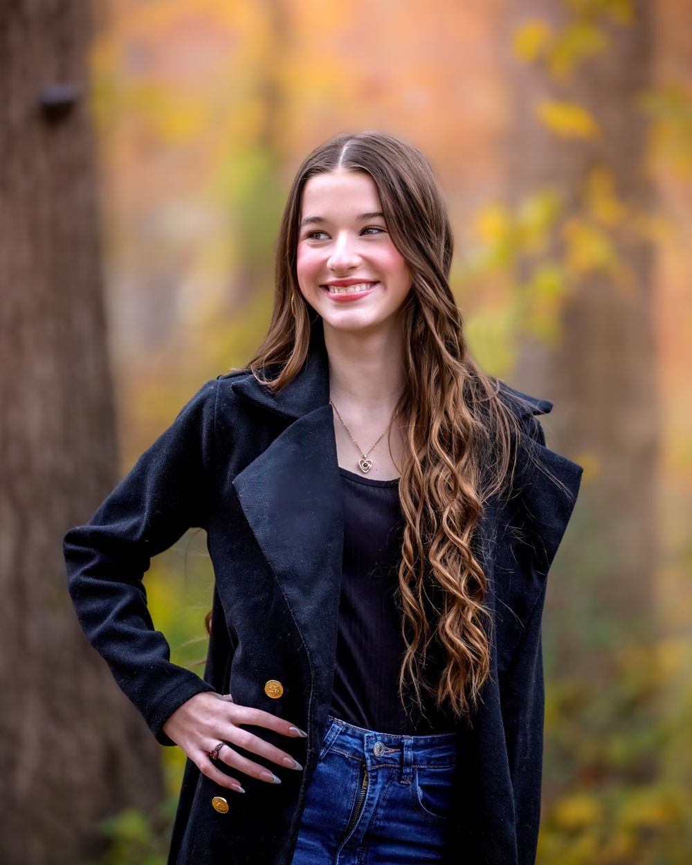 Outdoor autumn portrait showing someone in a black blazer and jeans with long wavy hair against blurred fall foliage.