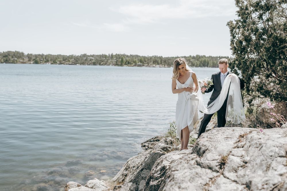 Brudpar går på en klippa vid havet, under ett bröllop på Djurö, Stockholm. Brudgummen håller brudslöjan i handen. Fotograf är Veronica Eng.