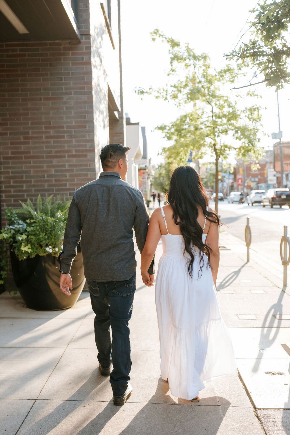A couple walking hand in hand down a sunny sidewalk, with the woman wearing a flowing white dress.