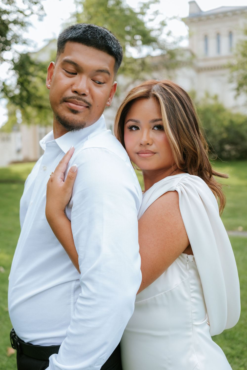 A couple in white attire poses outdoors near a building, the woman resting her hand on the man's shoulder.