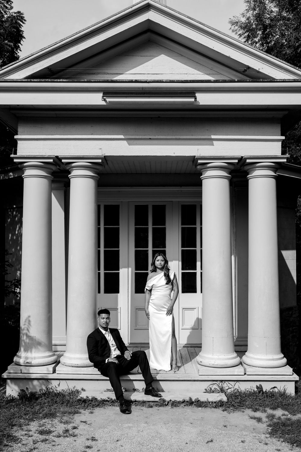 Black and white photo of a couple in formal wear posing on the steps of a classical building with tall columns.