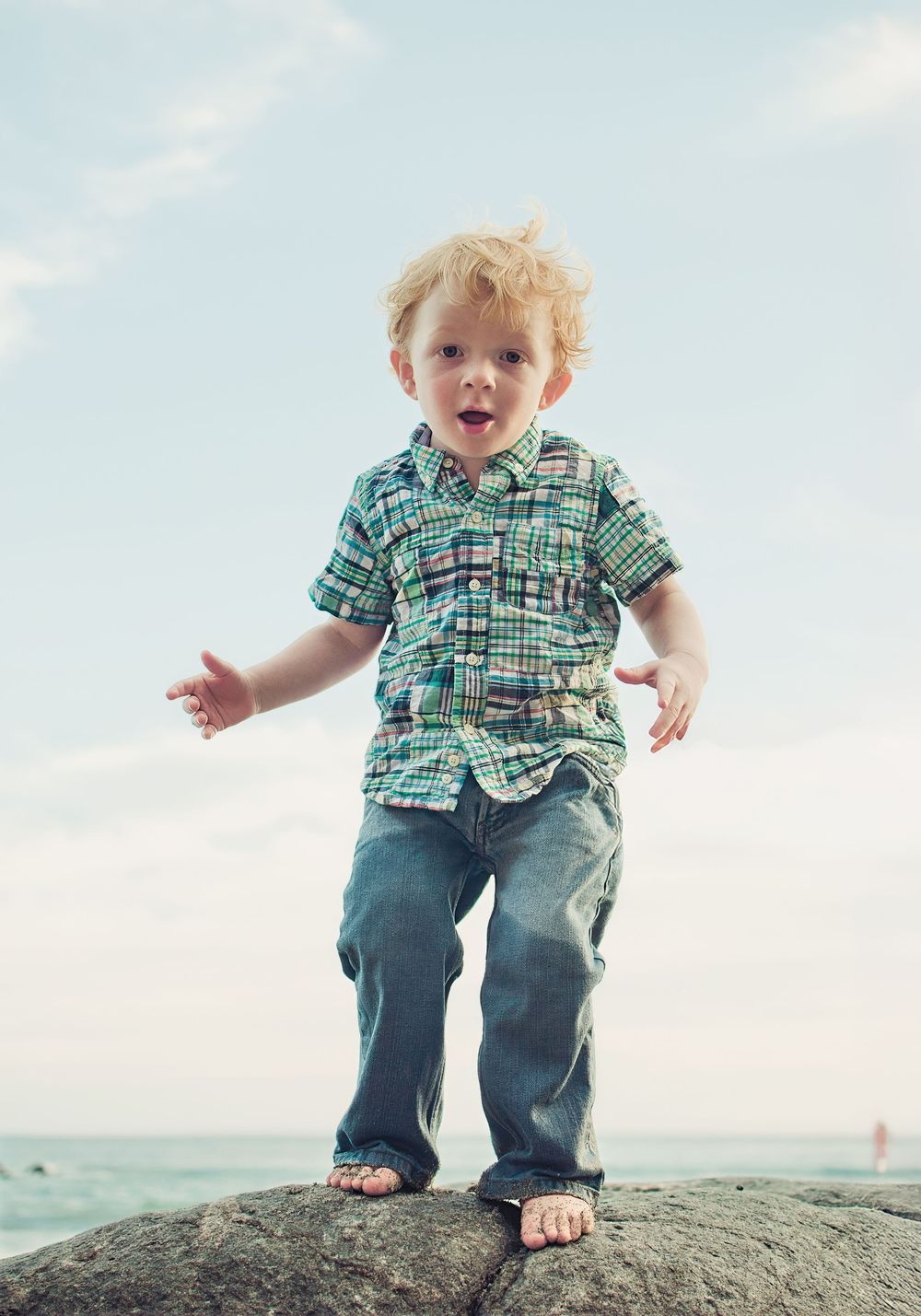 Curly red-haired toddler boy in plaid shirt and jeans standing barefoot on rocks near the ocean.
