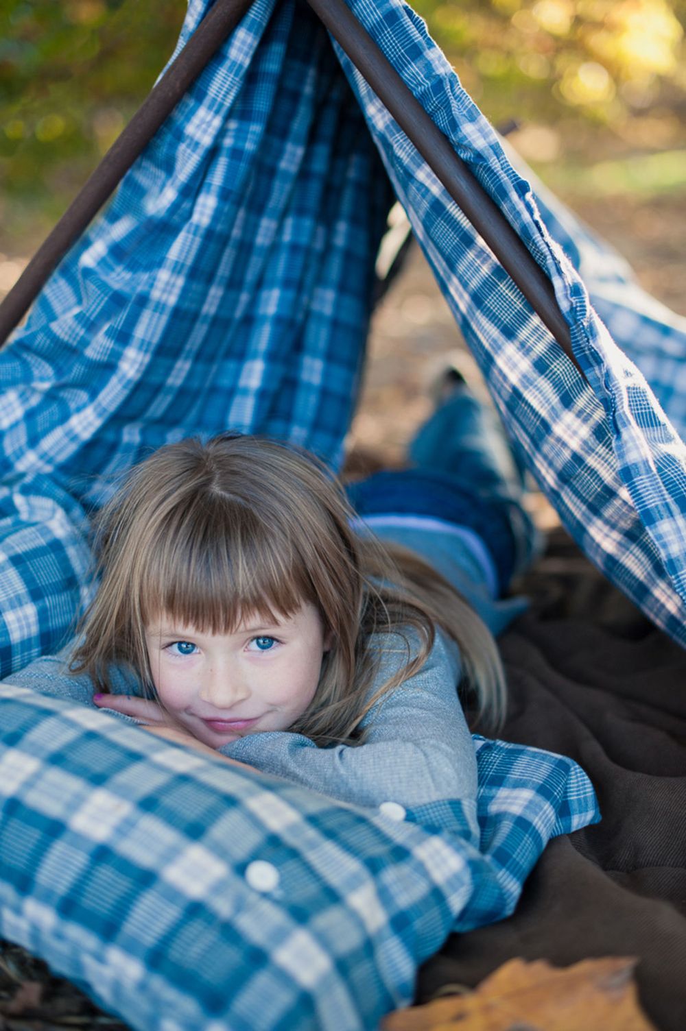 Young girl with blue eyes lying in a blue plaid tent outdoors, resting her chin on her hands.