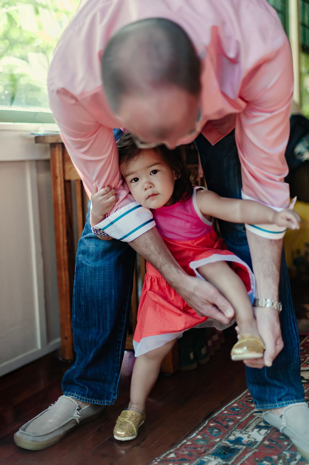 A father helps his toddler daughter put on gold shoes while she wears a bright pink dress indoors.