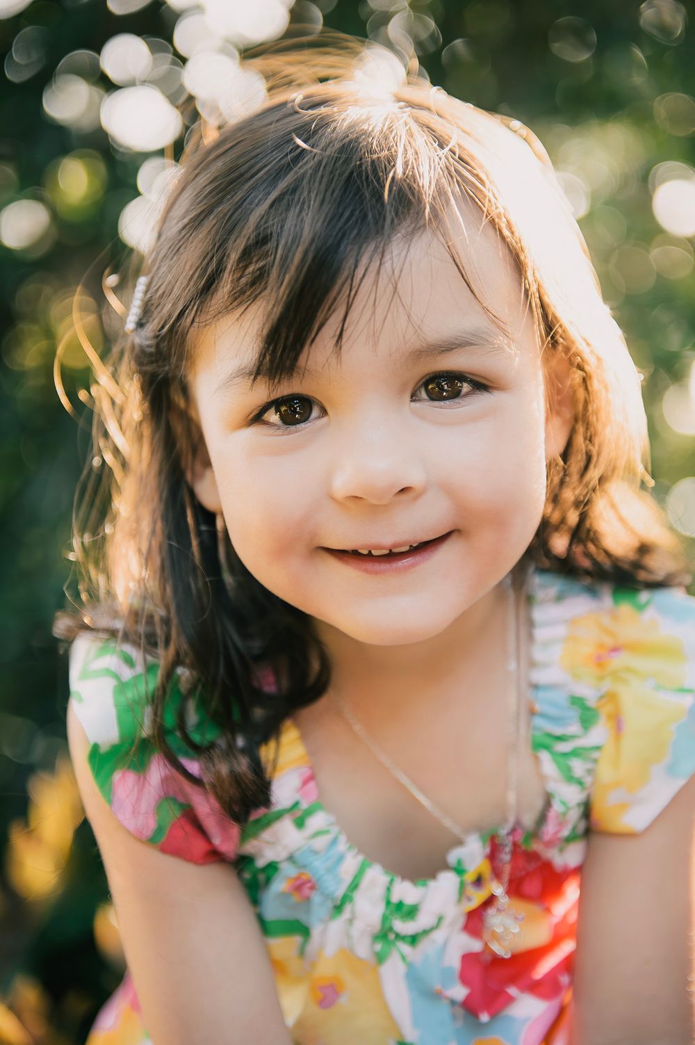 A smiling young girl with dark hair wearing a colorful floral dress poses outdoors in warm sunlight.