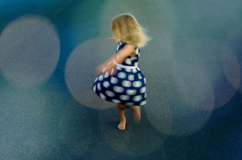 A young girl in a blue polka dot dress twirls barefoot on a colorful bokeh-lit floor.