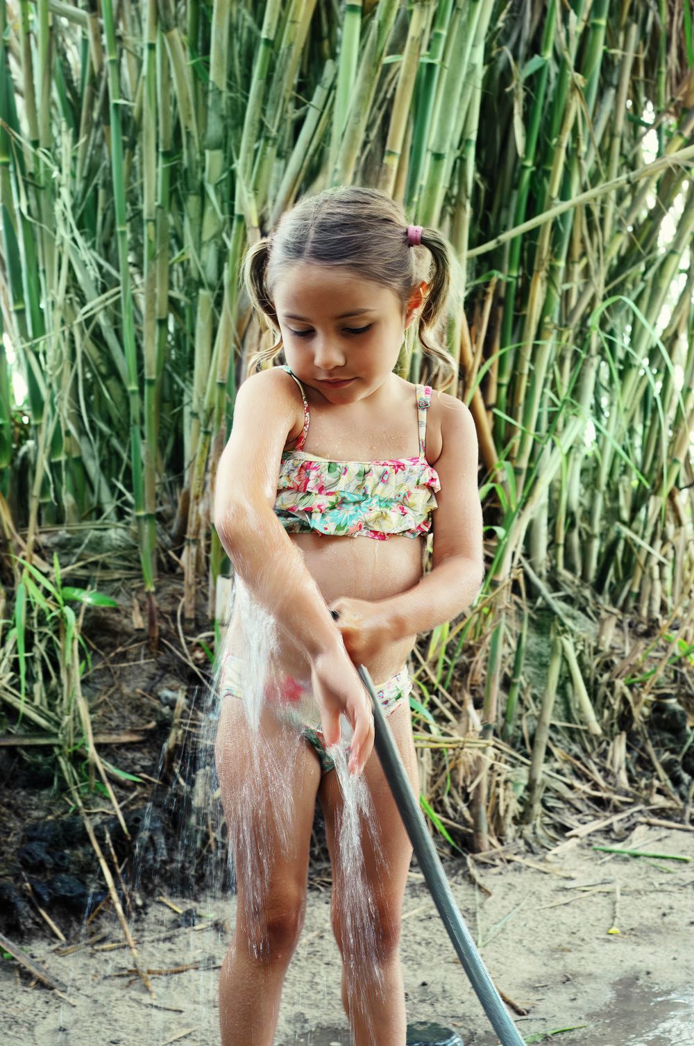 A young girl in a floral bikini plays with a garden hose near tall green bamboo plants outdoors.