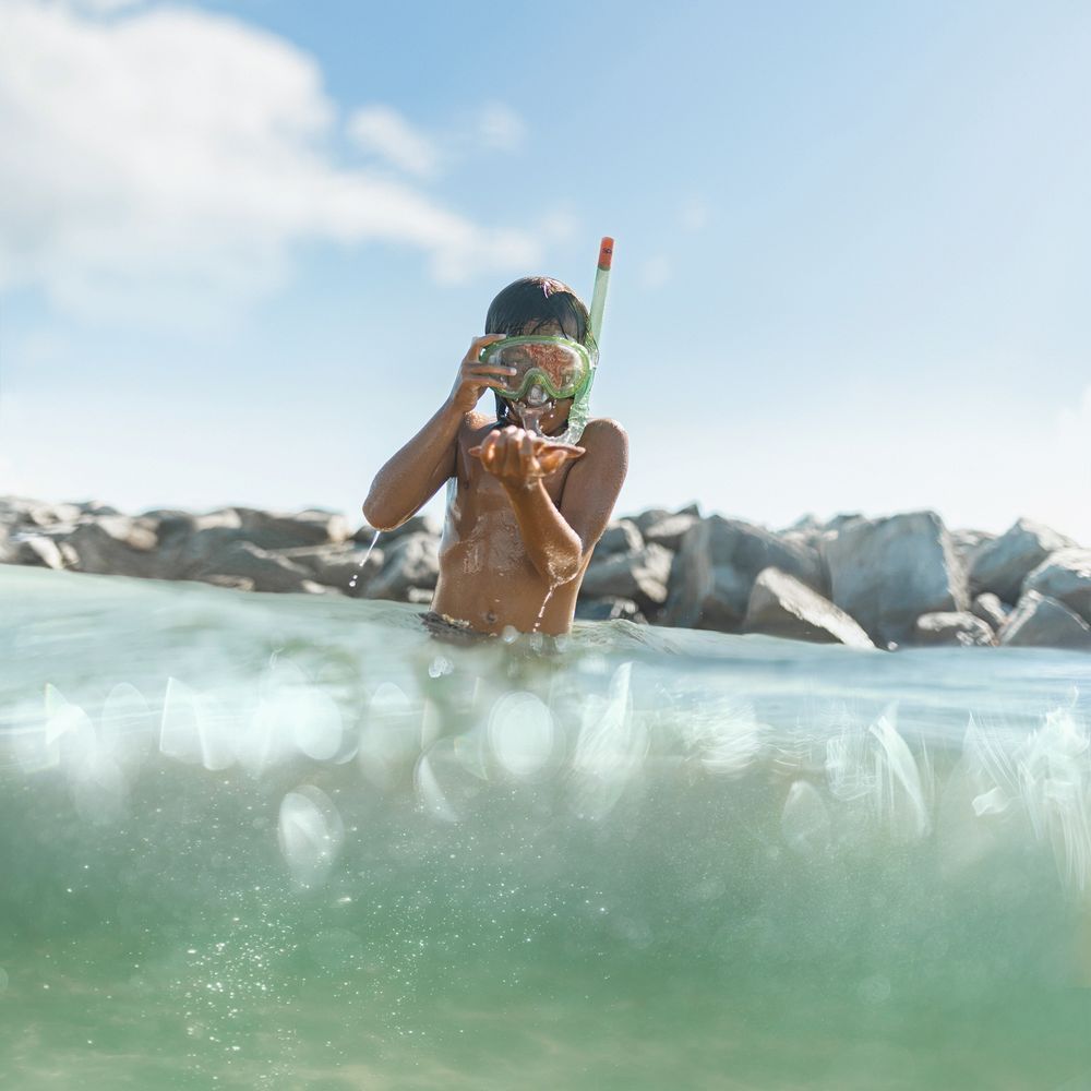 A child adjusting a snorkel mask at the ocean surface with rocky shoreline in the background.