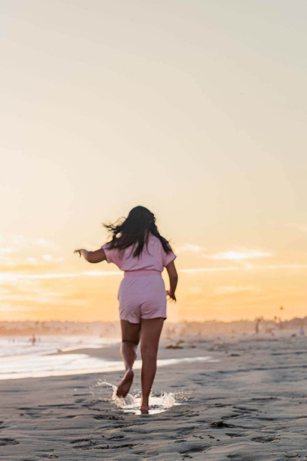 A young woman in a pink romper runs barefoot on a sandy beach at golden sunset.