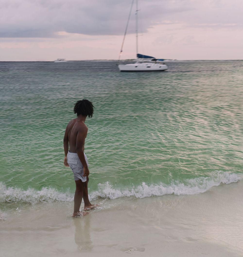A shirtless man in white shorts stands at the shoreline of a tropical beach with a sailboat anchored in calm turquoise waters.