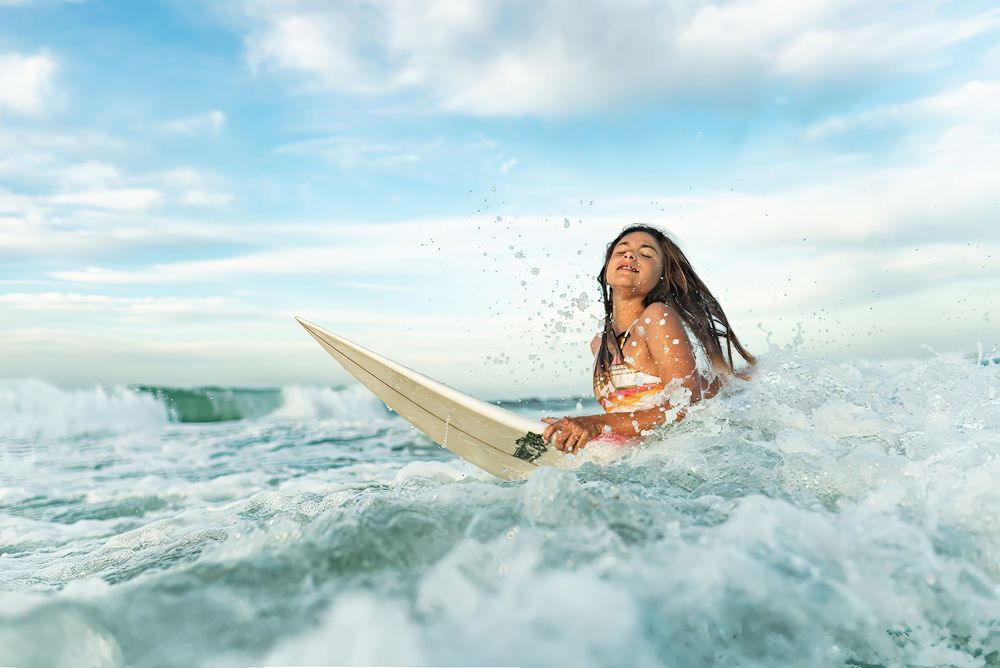 A young woman surfs ocean waves on a white surfboard under a partly cloudy blue sky.