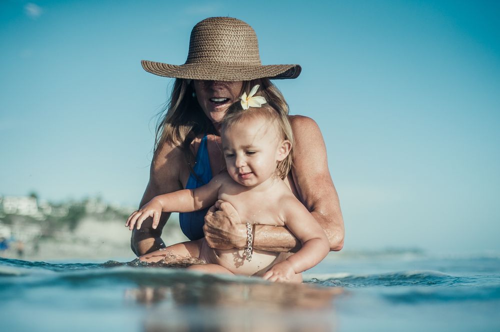A mother in a sun hat plays with her happy baby in shallow ocean waves on a sunny beach day.