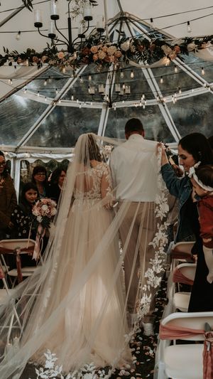 A wedding ceremony takes place under a clear tent decorated with twinkling lights and floral arrangements.