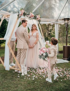Wedding party in soft beige and blush tones poses under elegant white draped outdoor canopy with scattered rose petals.