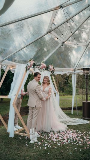 Romantic wedding ceremony under a clear tent with white drapes and scattered rose petals on the grass.