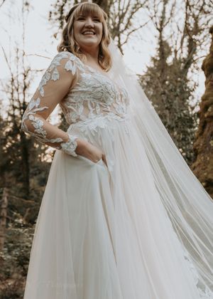 A bride in a flowing white wedding dress with lace details poses in a natural outdoor setting during golden hour.