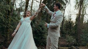 A couple in wedding attire dances together in a wooded outdoor setting during their wedding celebration.