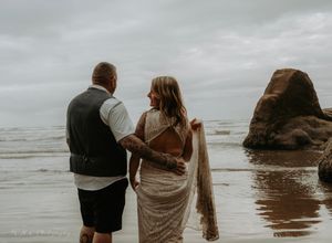 Two people stand together on a misty beach with large rocks visible in the ocean waves.