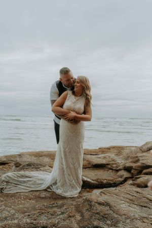 A romantic couple embraces on a rocky beach coastline with overcast skies in the background.