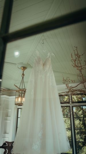 White wedding dress hanging from a rustic lantern in a sunlit room with white walls and windows.