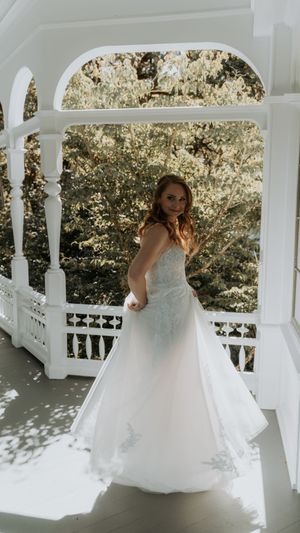 A bride in a flowing white gown poses gracefully on a white gazebo porch surrounded by dappled sunlight and greenery.