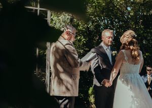 An outdoor wedding ceremony takes place in a sunlit garden with dappled shadows falling across the participants.