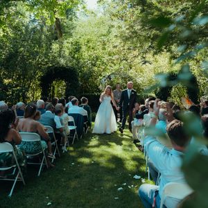 An outdoor garden wedding ceremony with guests seated on folding chairs beneath a floral arch on a sunny day.