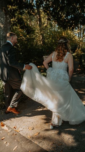 Wedding dress billowing in the breeze as couple walks along a sunlit autumn path with fallen leaves.
