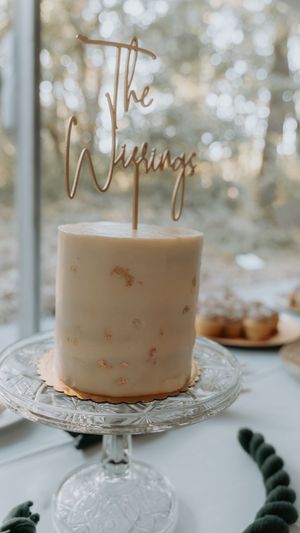 A simple white wedding cake with gold flecks on a crystal cake stand features a gold 'The Weddings' topper.