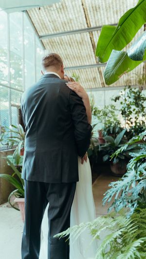 Newlyweds embrace in a greenhouse filled with tropical plants and natural light.