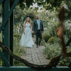 A wedding couple walks along a brick garden path framed by lush greenery and flowers.