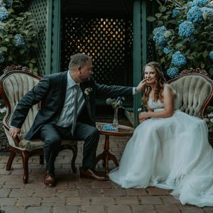 Couple shares tender moments on vintage furniture surrounded by blue hydrangeas in a garden setting.