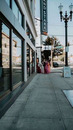 People in formal dresses stand outside a storefront on an urban street with ornate lampposts.