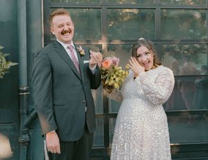 A joyful wedding couple poses against an industrial backdrop with green windows.
