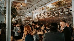 Wedding reception guests mingle at a rustic venue with exposed brick walls and string lights.