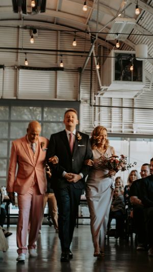 Wedding party members walk and laugh together in an industrial-style venue with exposed ceiling and string lights.