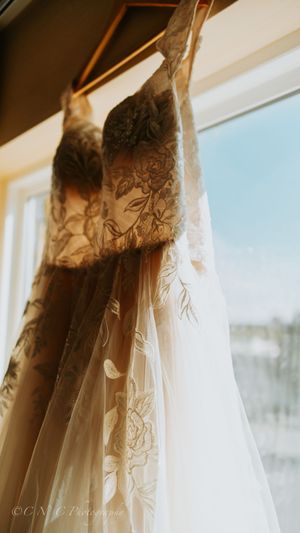 Elegant wedding dresses with delicate floral patterns hang in a row by sunlit windows.