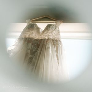 A delicate white wedding dress with lace details and tulle skirt hangs elegantly on a wooden hanger against a light wall.