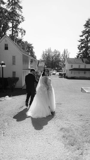 A wedding couple walks across a grassy field with white buildings in the background on their special day.